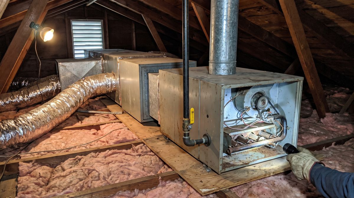 Horizontal gas furnace with its panel off in a tract-house attic, coil and ducts attached, a flue rising through the roof and a flashlight on the burners